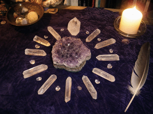 A healing-table crystal grid: clear quartz points arranged in a circle around a large amethyst, on a purple cloth, by candlelight.
